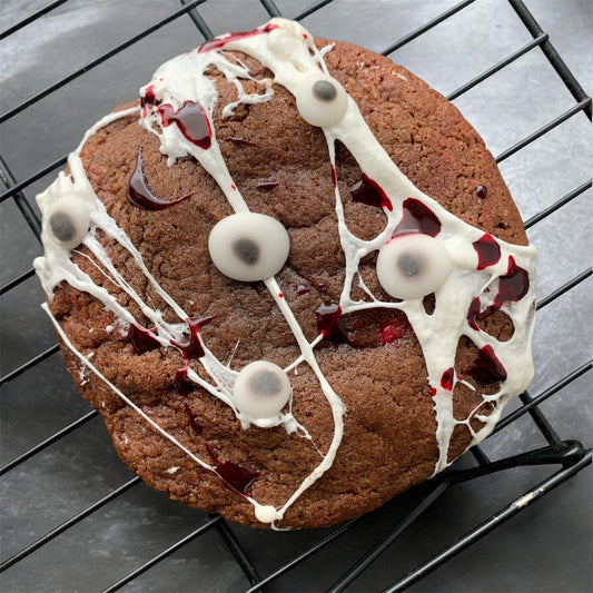 A large chocolate chip cookie decorated with white drizzle, dark red syrup, and gummy eyeballs, resting on a black wire cooling rack over a dark surface.