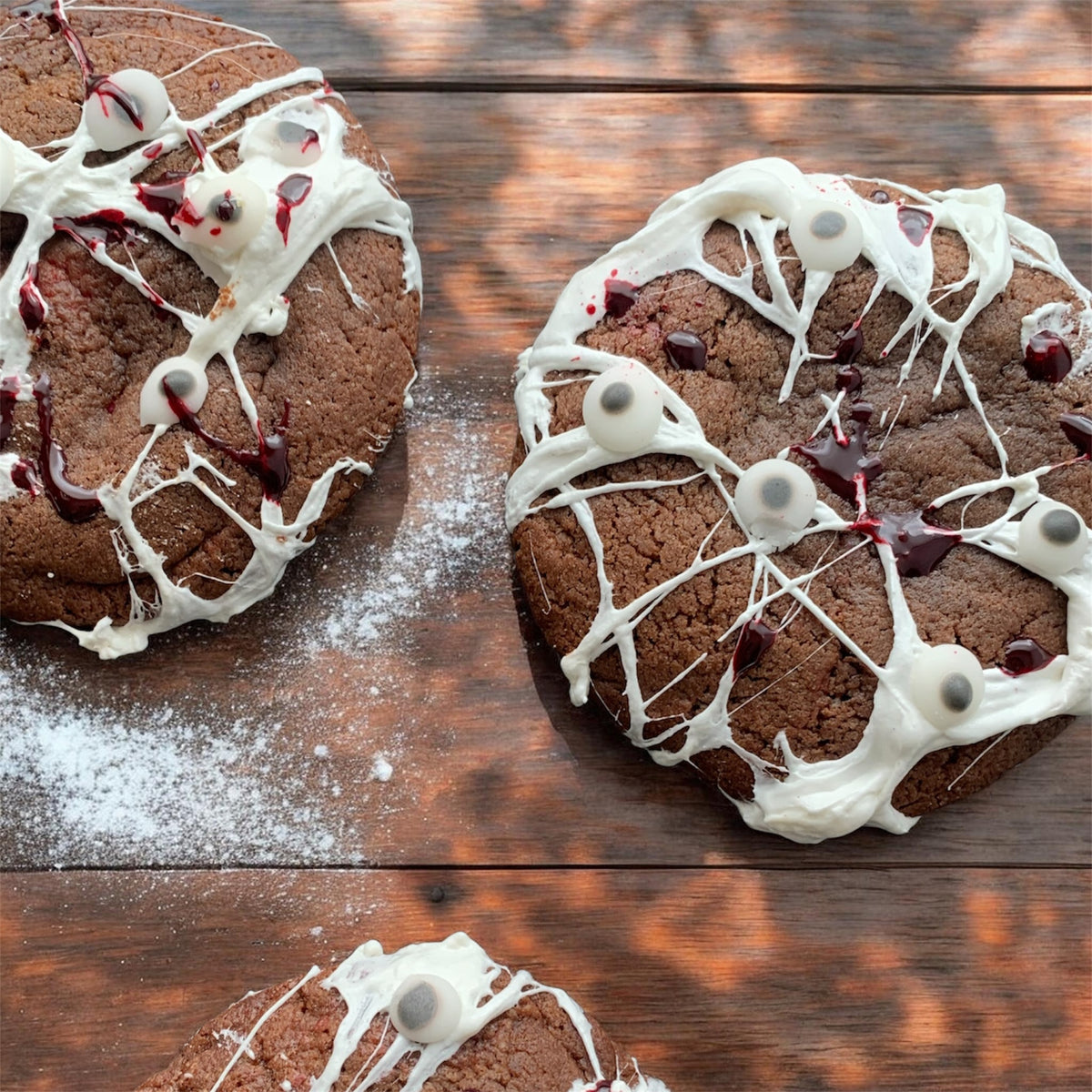 Three chocolate chip cookies decorated with white icing, dark red drizzle, and gummy eyeballs, arranged on a rustic wooden surface with a dusting of powdered sugar.
