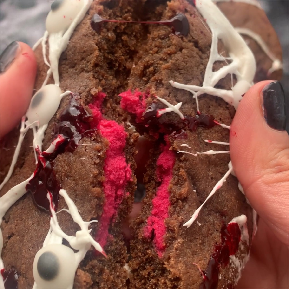 A close-up of a chocolate cake or cookie being held by two hands, broken open to reveal bright pink-red filling, with white and dark red drizzle on top.